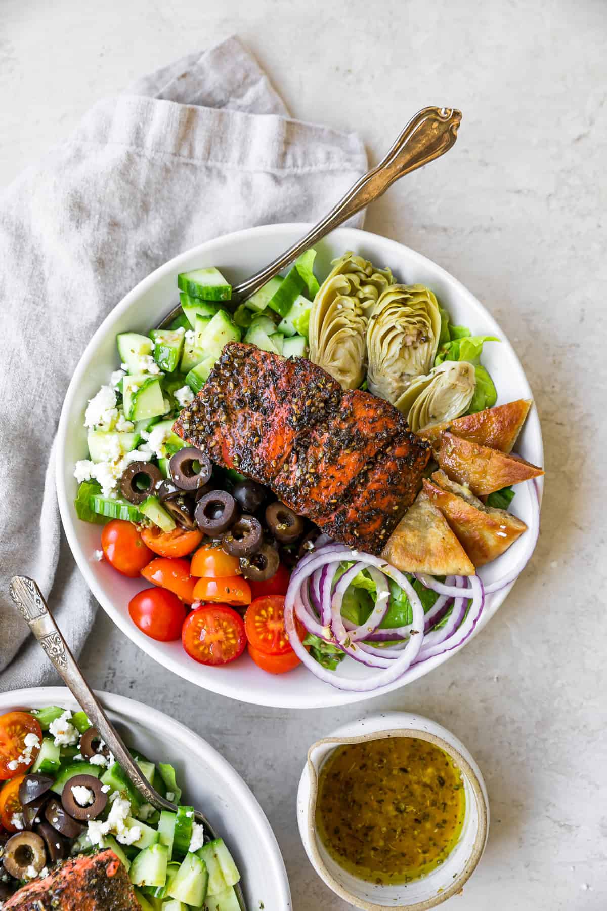 greek salmon salad in bowls with fork next to linen and dressing bowl