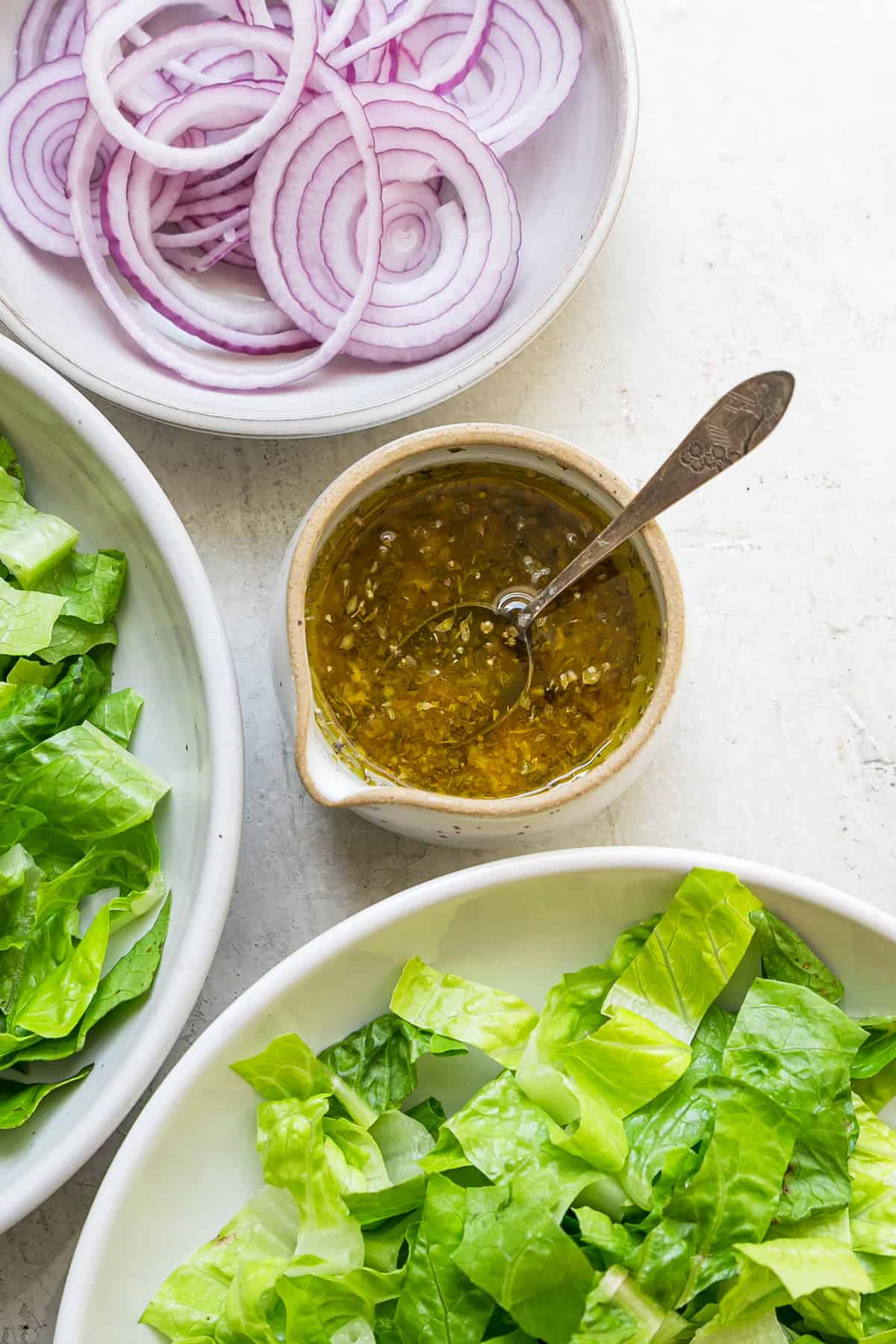 overhead view of homemade greek salad dressing in small dish next to bowls or romaine and red onion rings