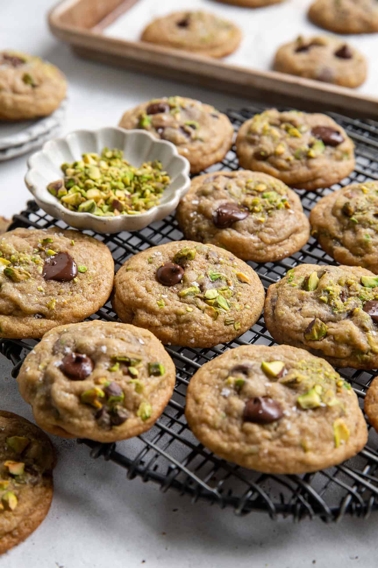 pistachio chocolate chip cookies on black wire rack with cookies on pan in background.