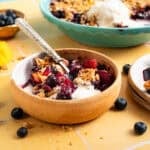 mango blueberry crisp in pottery bowl on yellow tile next to teal pie dish.