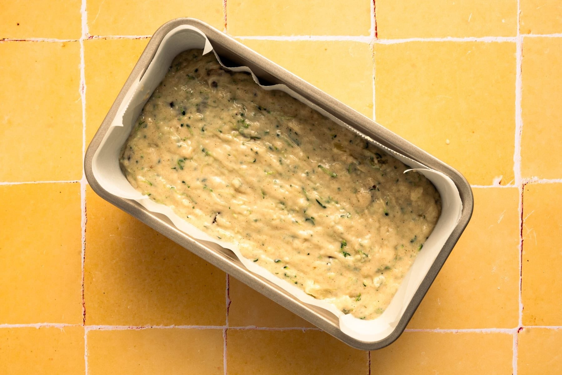 zucchini bread batter in parchment lined loaf pan on yellow tile.