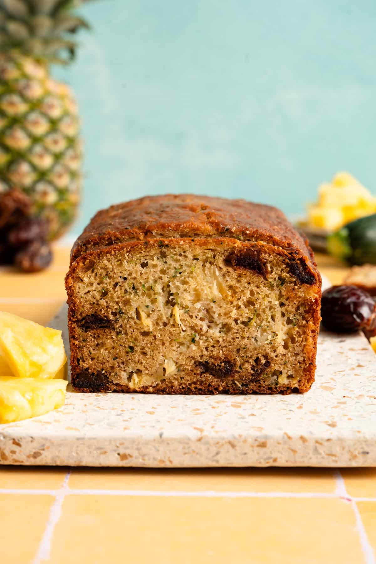 zucchini pineapple bread recipe on yellow tile with turquoise background, next to fresh pineapple, dates and zucchini.