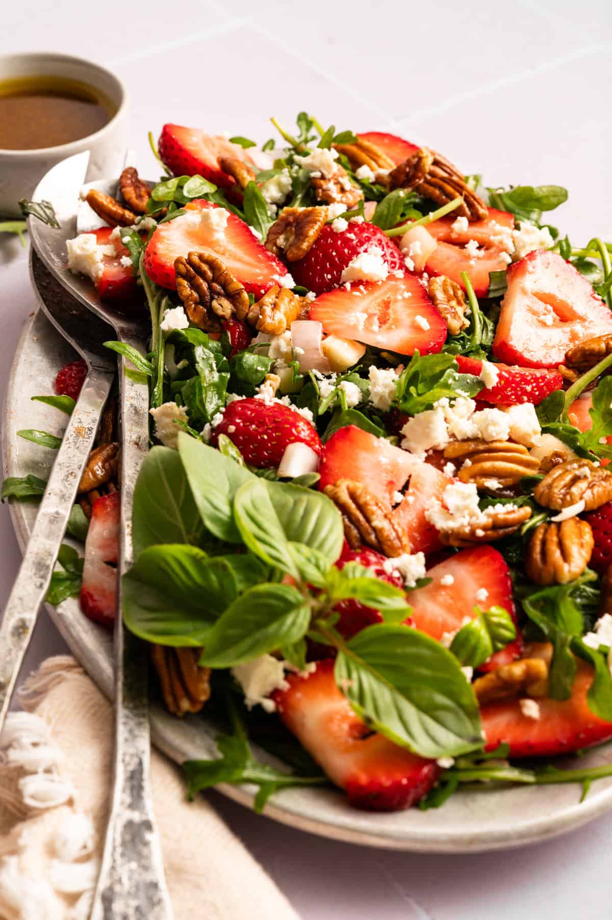 strawberry arugula salad with silver serving spoons on off-white tile background.