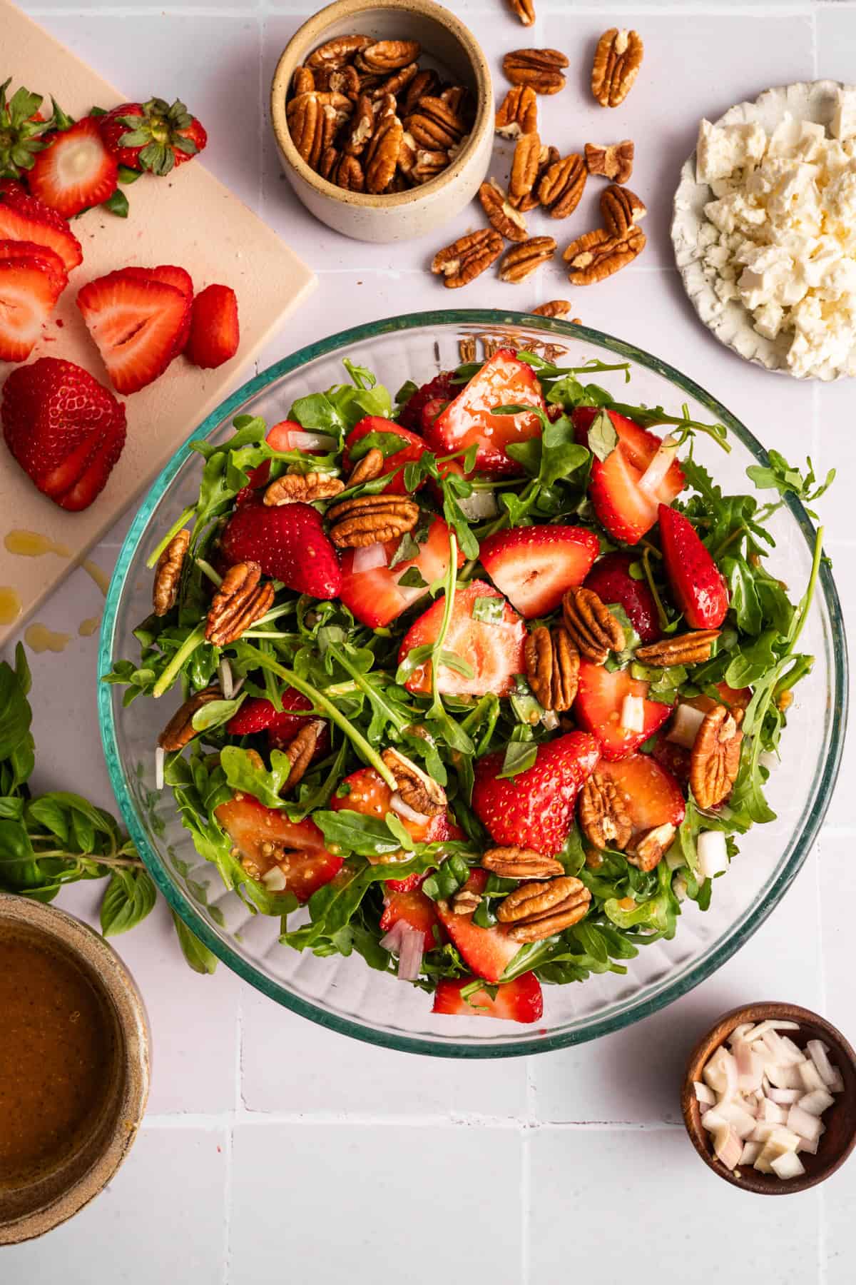 overhead view of strawberries and pecans in a glass salad bowl, next to feta, pecans, shallots and dressing.