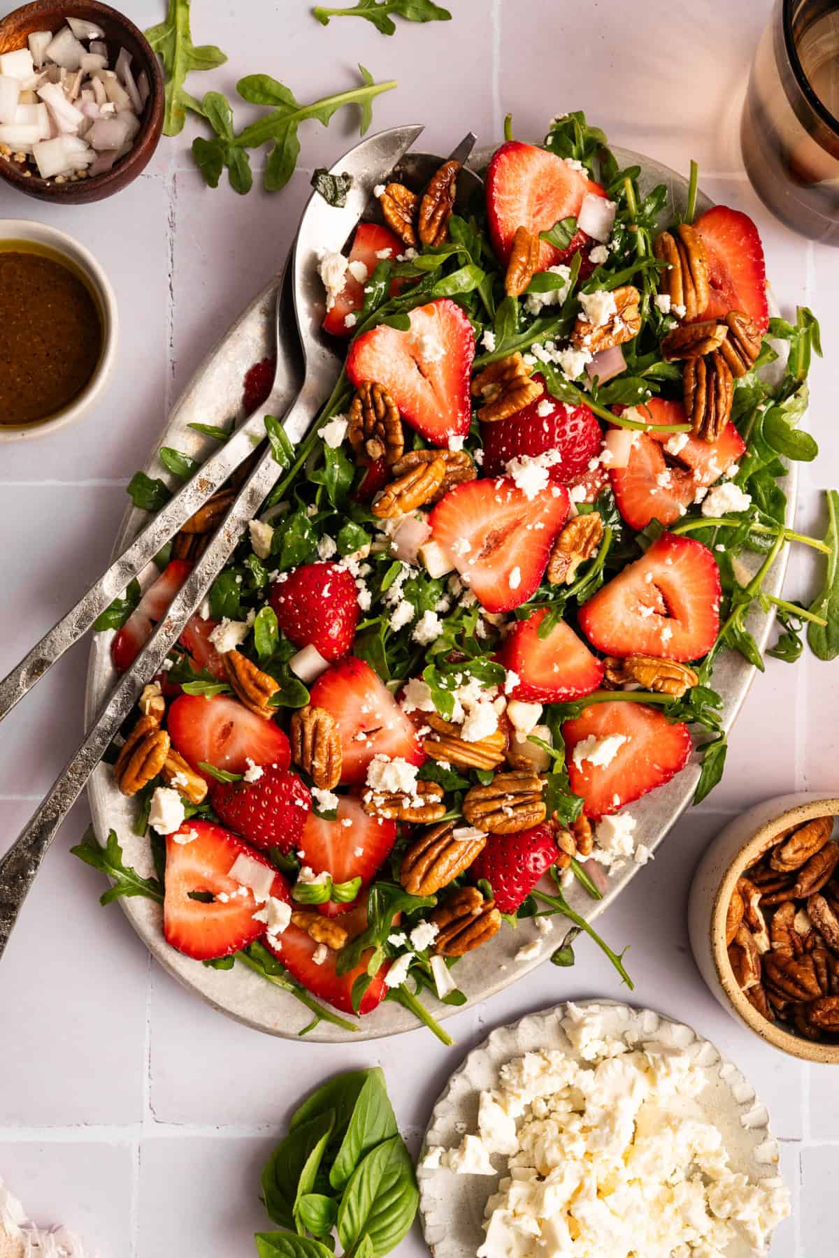 overhead view of strawberry arugula salad with feta and pecans, next to small bowls of ingredients and glass of water.