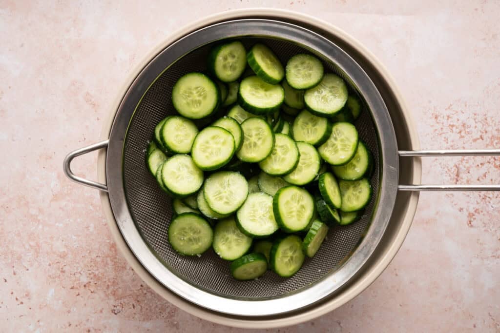 sliced mini cucumbers in metal strainer over bowl.