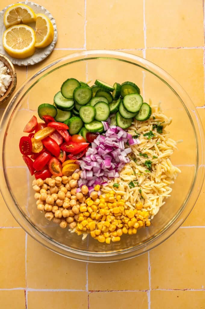 overhead view of cucumbers, tomatoes, corn, red onion, chickpeas and orzo in large bowl.