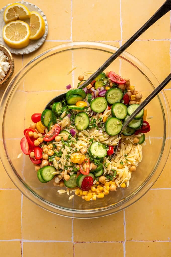 fresh vegetables with salad servers in glass bowl with orzo pasta.