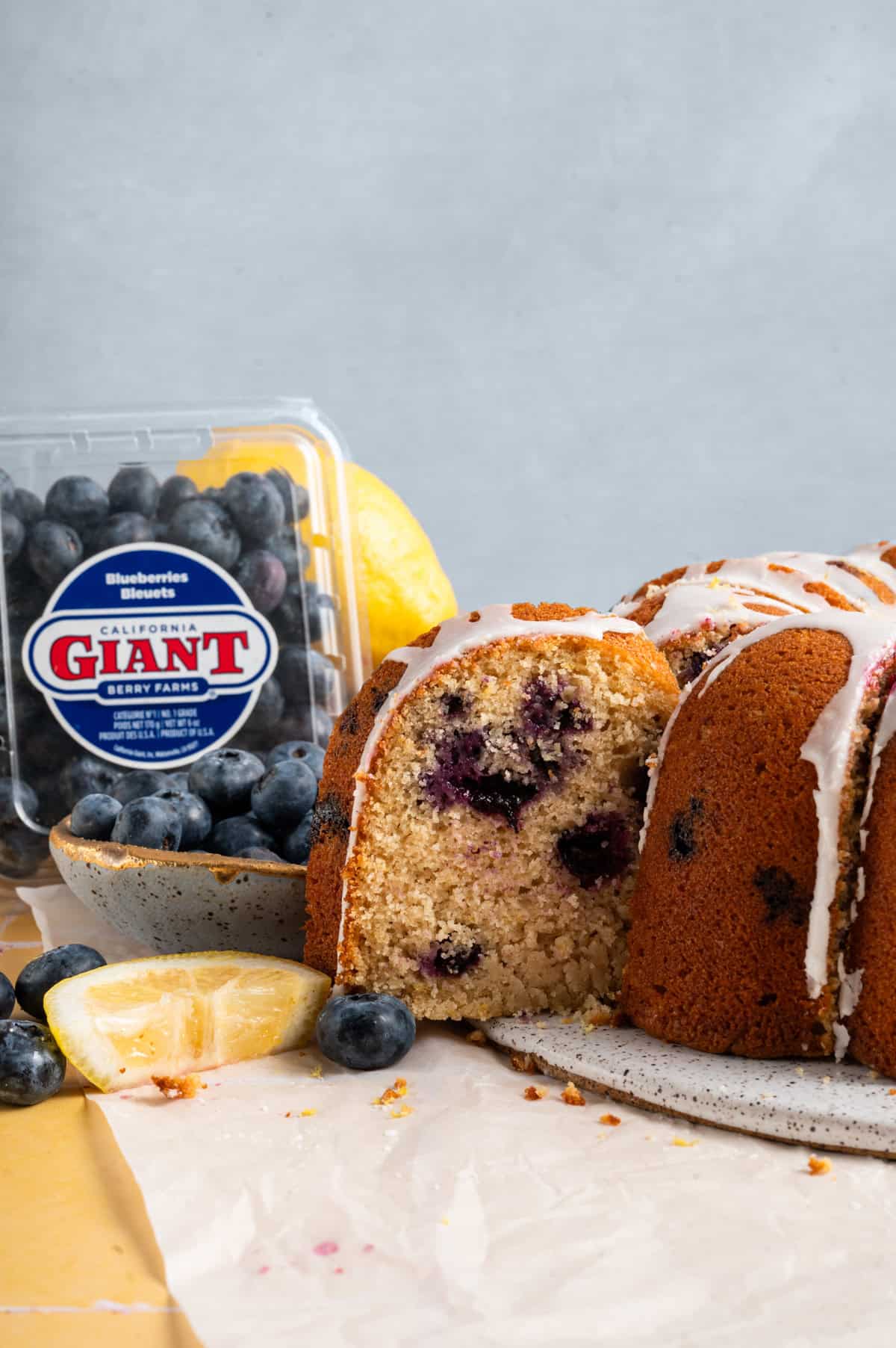 blueberry lemon bundt cake on round platter next to blueberry clamshell in background.