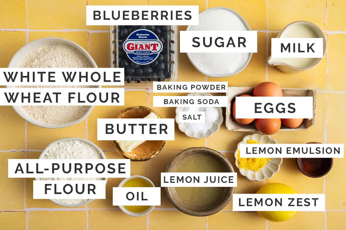The image shows various baking ingredients for blueberry lemon bundt cake, labeled with their names, arranged on a yellow tiled surface.