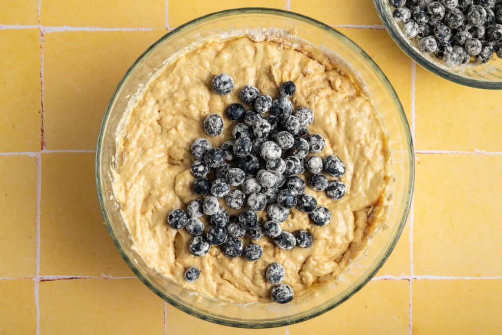 glass bowl filled with flour dusted blueberries on top of lemon cake batter.