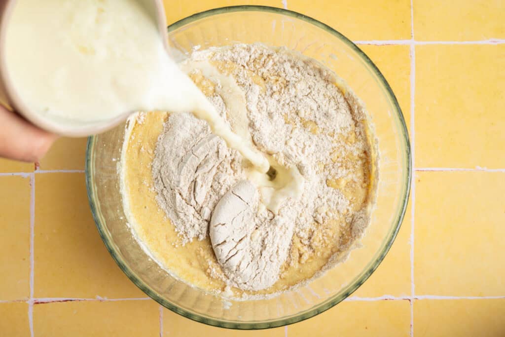 Pouring milk into a bowl with flour on yellow tile surface.