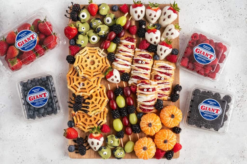 wooden board with healthy halloween snacks like chocolate covered berries, hand pies, spider waffles, clementines and california giant berries.